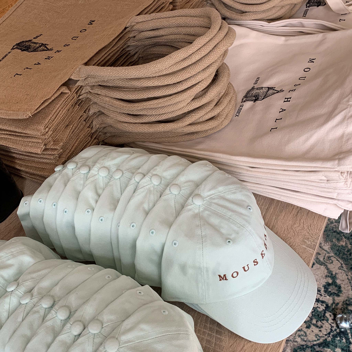 an image showing a bulk order for a company called Mousehall. The image shows mint green caps, jute shoppers and totes all embroidered with the corporate logos.
